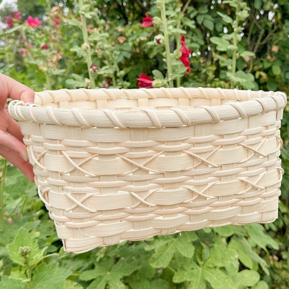 Woman holding a cross-stitch napkin basket sitting against a backdrop of hollyhocks.
