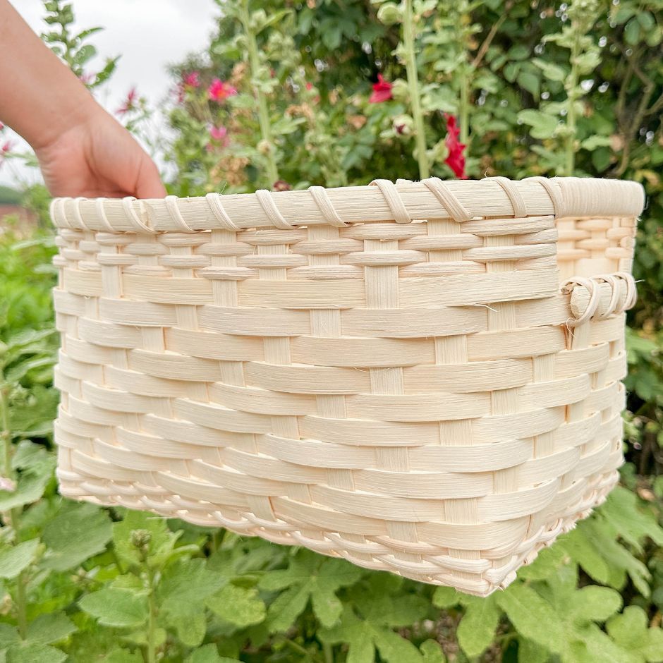 A woman's hand holding a handled storage basket against a background of hollyhocks.