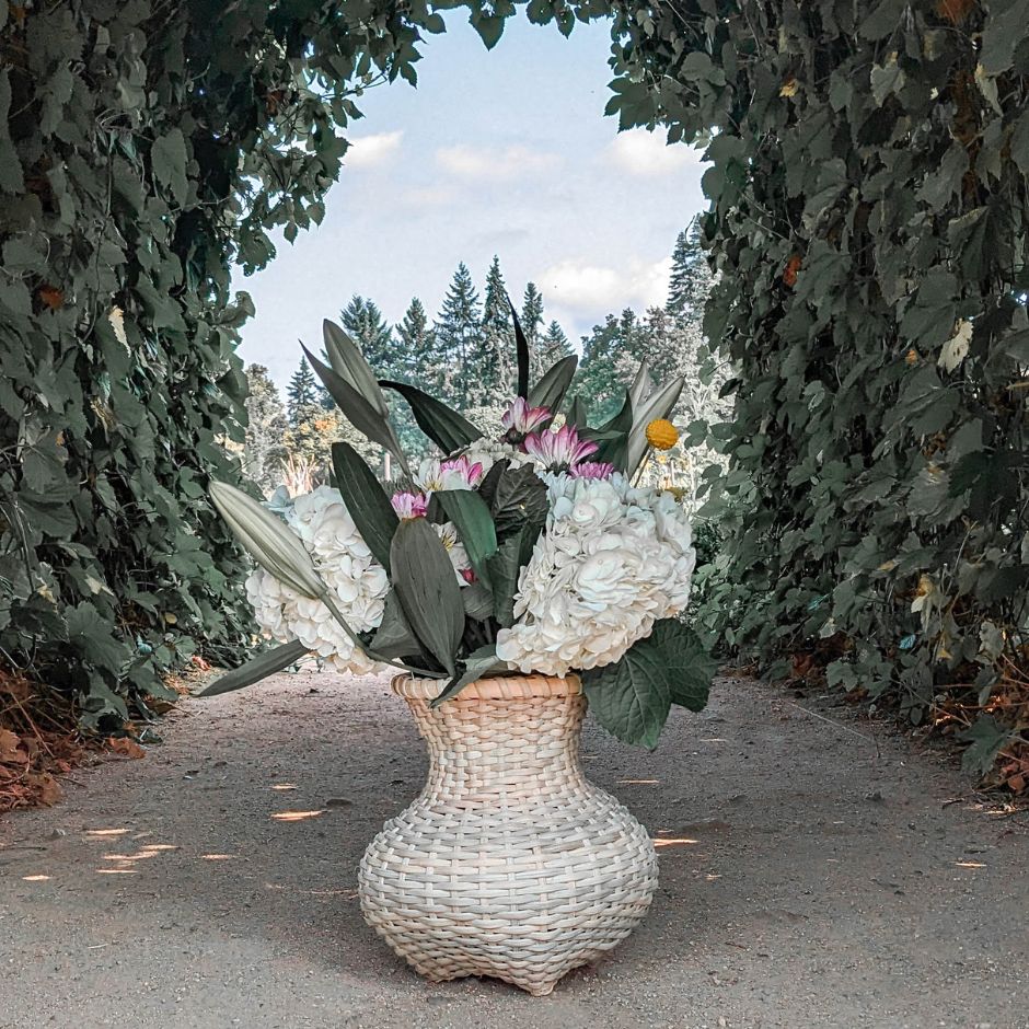 A vase shaped basket weave basket sits under an arch of hops vines. the basket is stuffed with white hydrangea and lily flower arrangement.