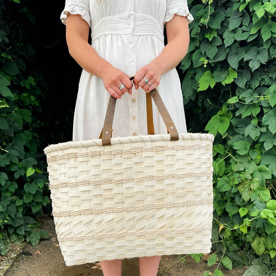 Person holding a woven basket with a white dress against a green leafy background.