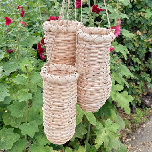 Three hanging Flower Vase Baskets against a backdrop of hollyhocks.