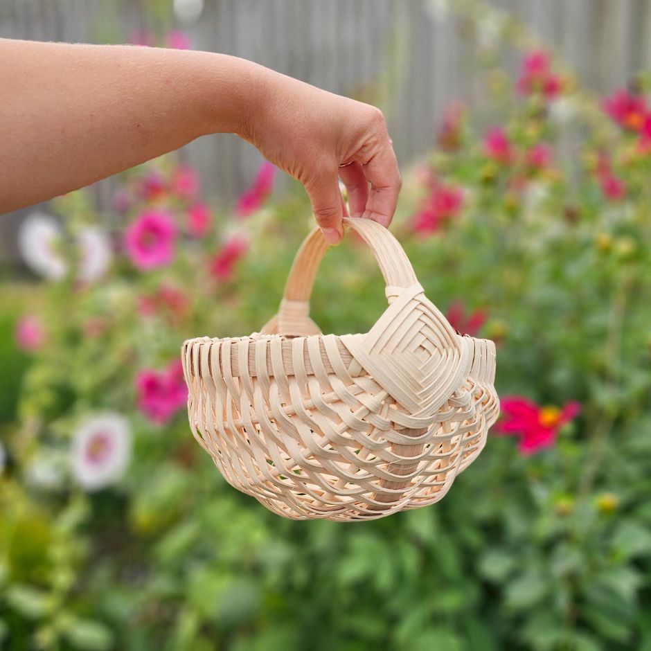 Woman's hand holding a melon basket by the handle. A garden of cosmos and hollyhocks sits in the background. 