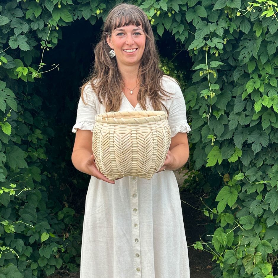 Woman holding a woven catshead basket. She is framed by hop vines.