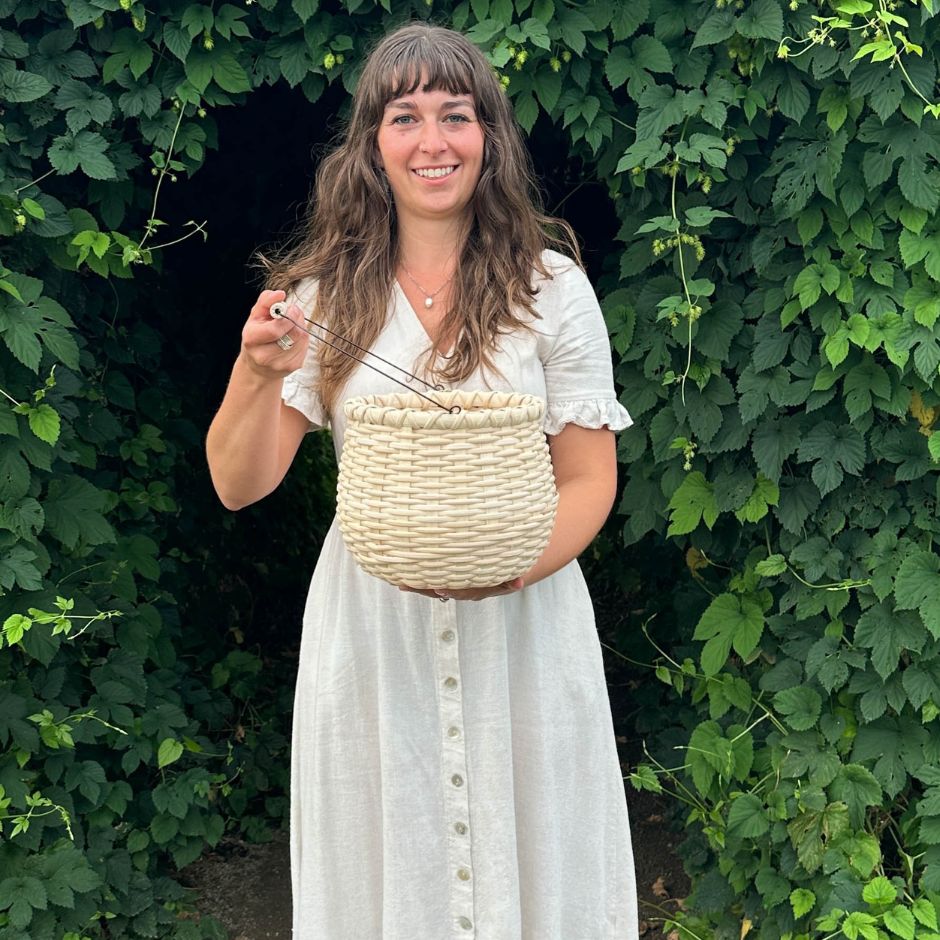 Woman in a cream linen dress holding a bean pot basket. She is framed by an arch of hop vines. 