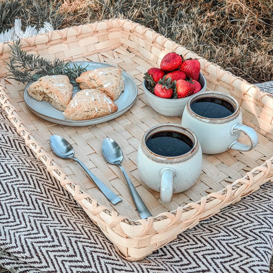 Coffee table basket sitting on a black and white blanket in the grass. The basket is set with two full coffee mugs, two teaspoons, a bowl of strawberries, and a plate of scones. 