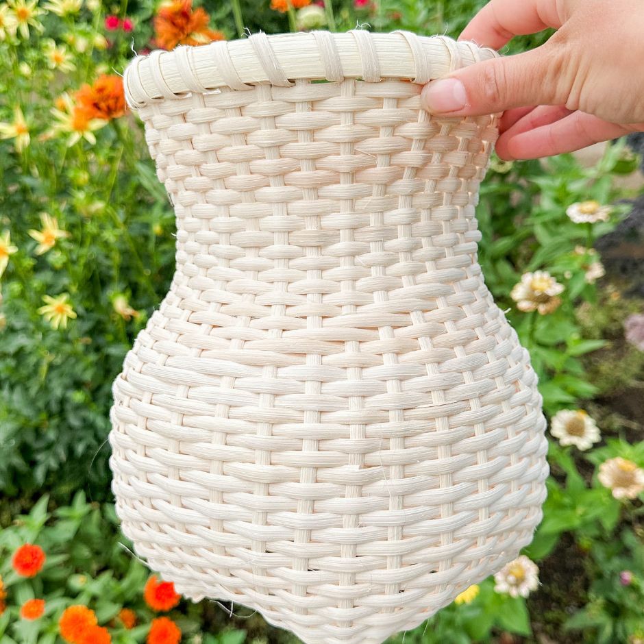 A woman's hand holds a vase shaped stair-step weave basket against a backdrop of marigolds. 