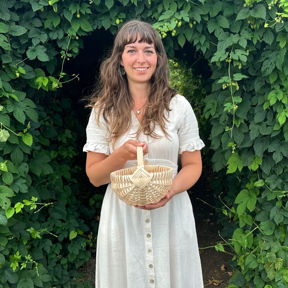 Woman in a cream linen dress holding a melon basket. She's framed by an arch of hop vines. 