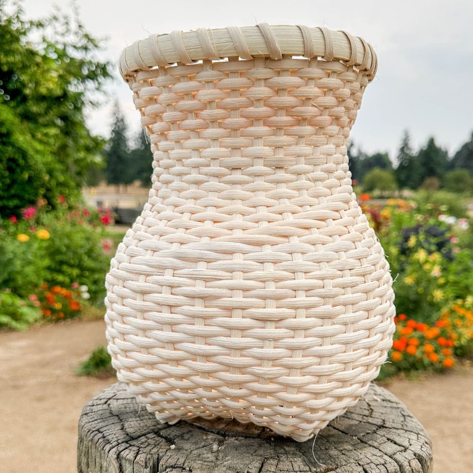 A vase shaped basket sits on a tree stump. A backdrop of bright flowers fills the background. 