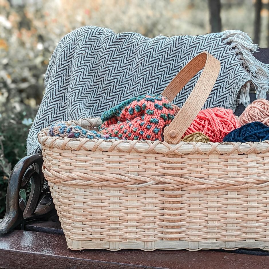 Braided market basket sitting on a garden bench. The basket is overflowing with a knitting project. A black and white blanket is draped over the bench in the background. 