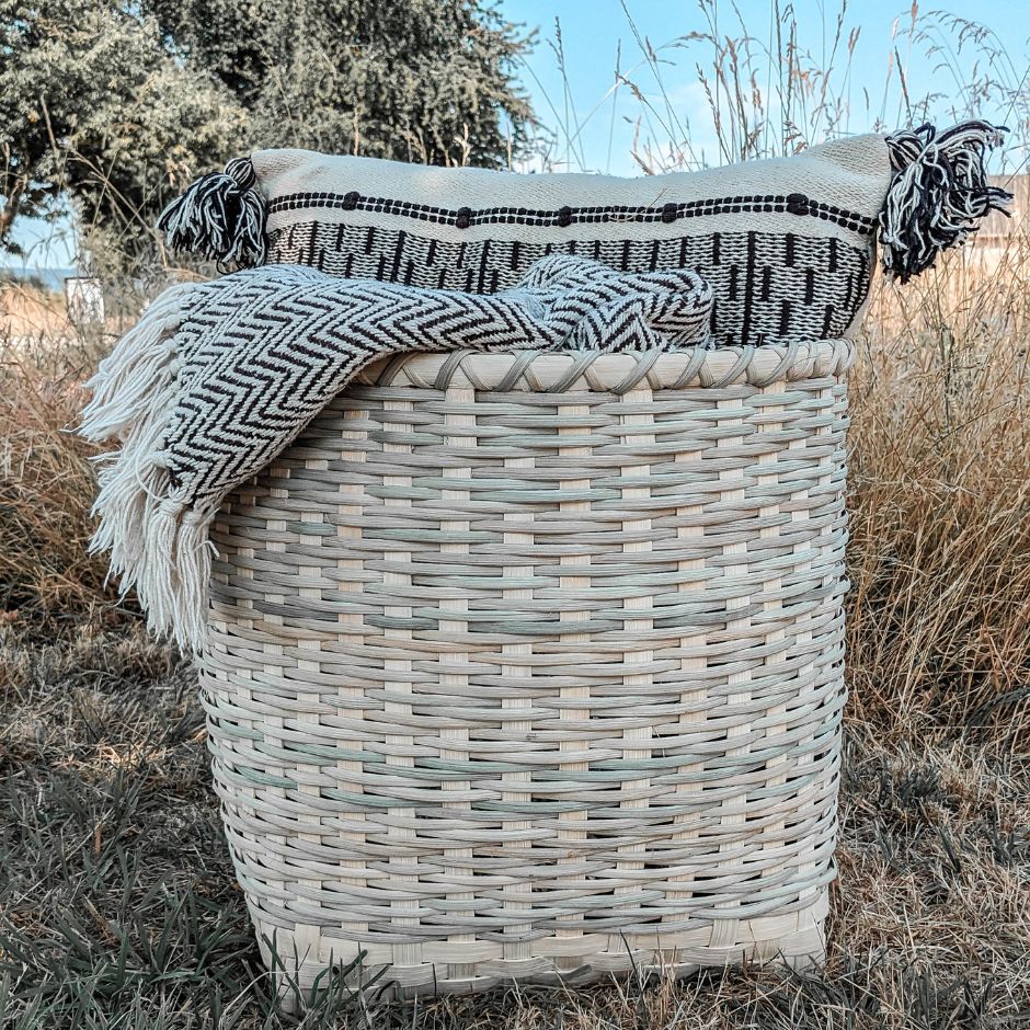 Twill blanket basket sitting in a grassy field. The basket holds a black and white pillow and blanket. 