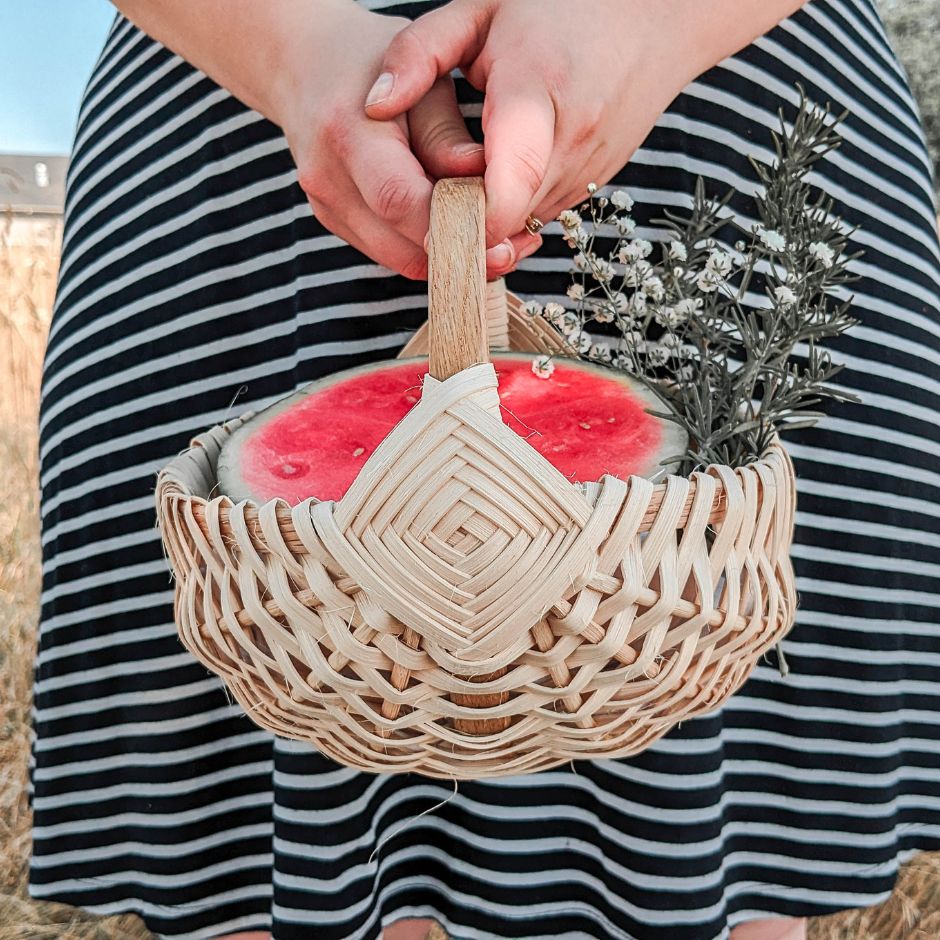A woman in a black and white striped skirt holds a melon basket filled with a cut watermelon and a sprig of baby's breath and rosemary. 