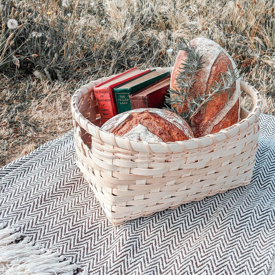 A handled storage basket sitting on a black and white blanket in the grass. The basket if filled with crusty loaves of bread and vintage books.