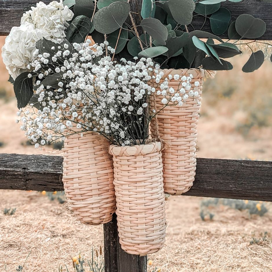 A trio of woven rattan vase baskets hangs from a split rail fence. The baskets are overflowing with baby's breath, white hydrangeas, and eucalyptus leaves. 