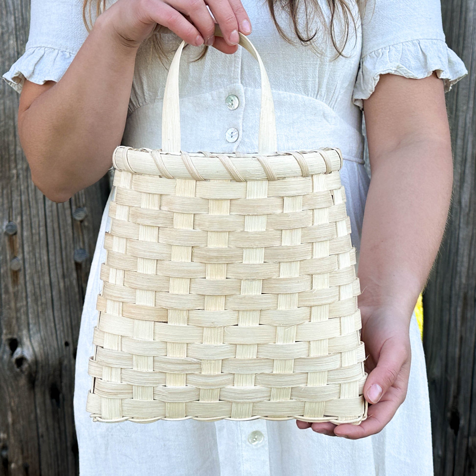 Person holding a woven handbag in front of a wooden fence