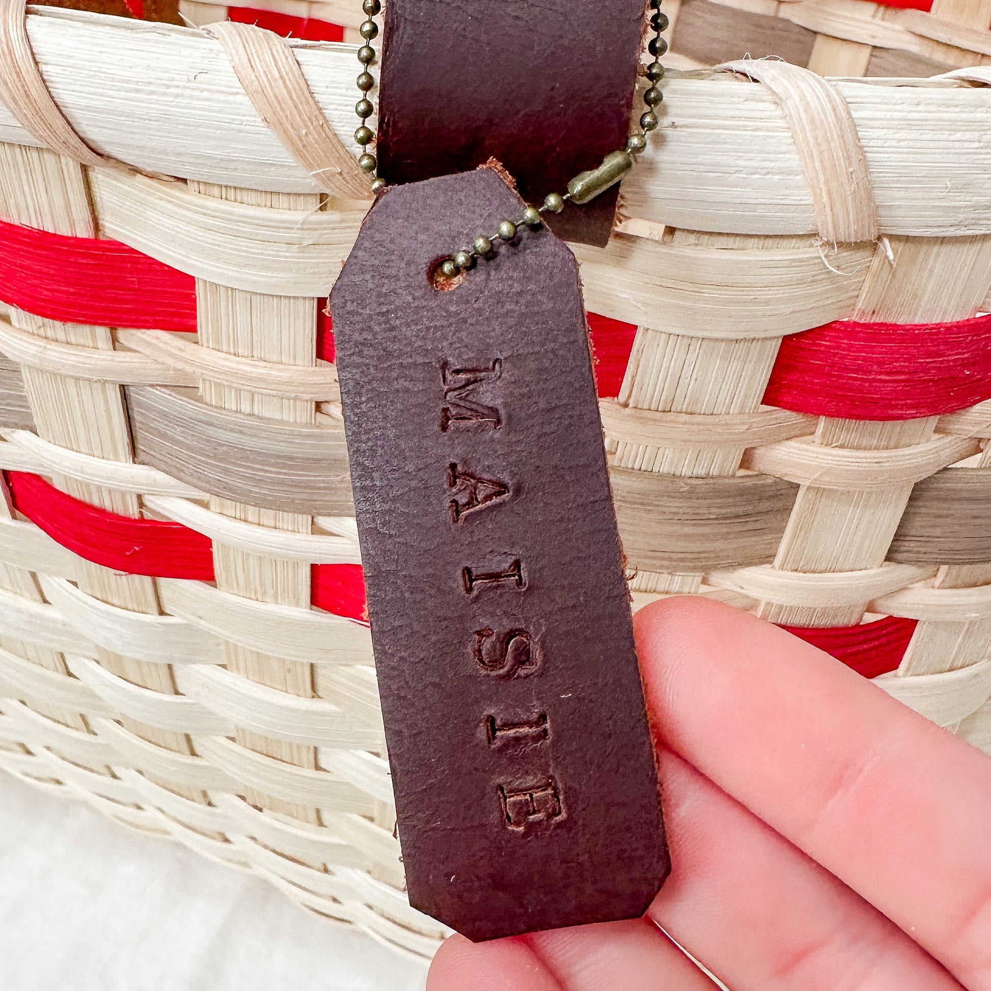 Hand holding a leather tag with 'MAISIE' engraved, against a background of a woven basket with red and beige stripes.