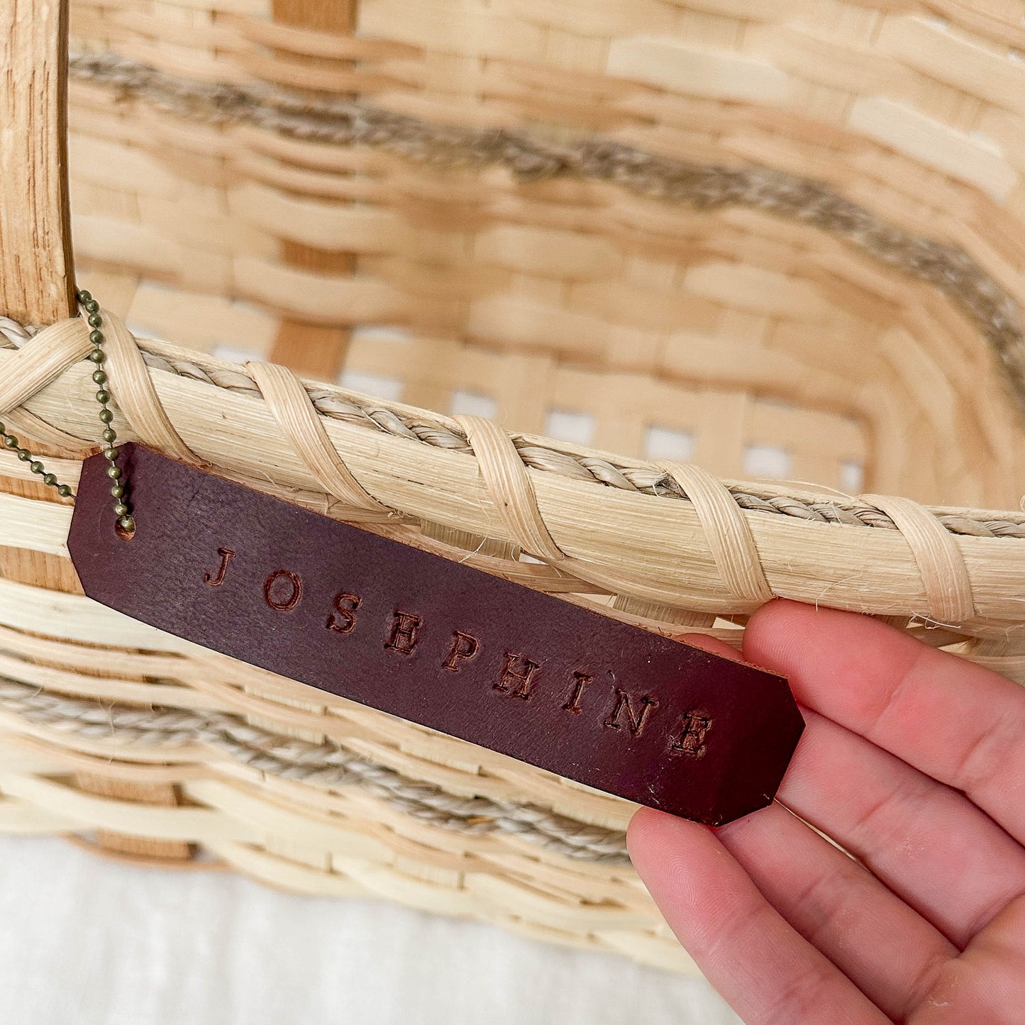 Hand holding a leather tag with 'Josephine' engraved, against a woven basket background.