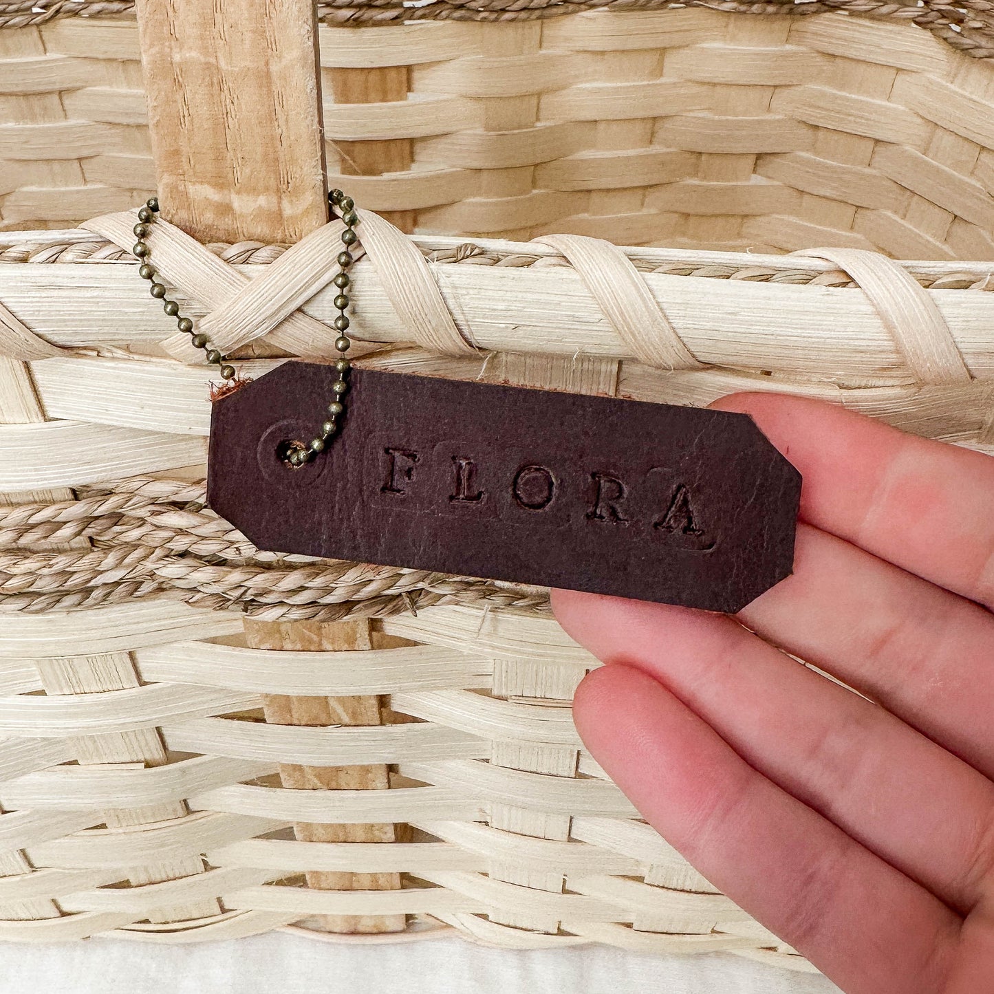 Hand holding a leather tag with 'FLORA' engraved, attached to a woven basket.