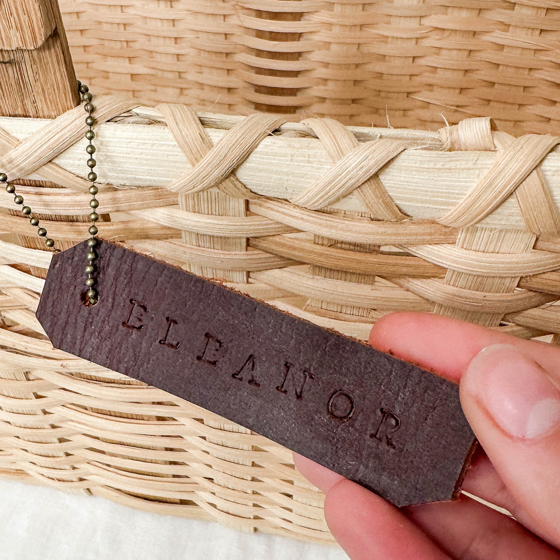 Person holding a leather tag with 'ELEANOR' engraved against a woven background